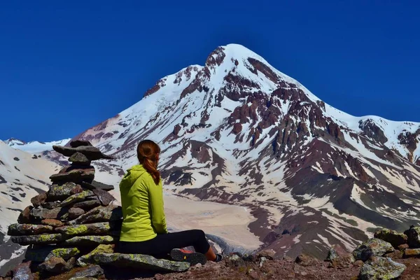 黄色のスウェットシャツを着た少女が石の男の隣に座って 氷河で覆われたカズベク山の頂上を見ている 山の中で美しい晴れた日の高 ジョージア — ストック写真