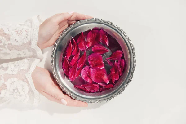 Kadın ellerinde koyu pembe şakayık yaprakları ve beyaz arka planda temiz suyla dolu bir kasede. Organik kozmetik, spa konsepti. Boşluğu kopyala, düz yat, üst görünüm.
