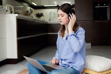 Kulaklıklı güzel bir kadın dizüstü bilgisayar kullanıyor, ekrana bakıyor, mutfakta duruyor, sosyal ağda mutlu genç bir kadın sohbet ediyor, online alışveriş yapıyor, çalışıyor, karantinada duruyor.