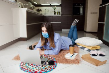 Tıbbi maskeli genç bir kadın evde dizüstü bilgisayar kullanarak başarılı bir şekilde çalışıyor. Ev karantinası, izolasyon, önleme COVID-19, sosyal mesafeler, ev ofisinden serbest çalışma, kendini izole etme