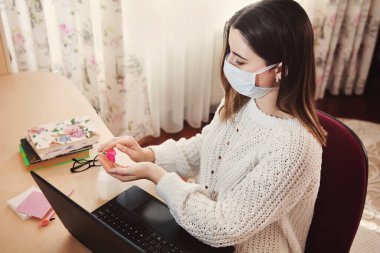 Coronavirus konsepti. Maskeli kadın evden çalışıyor. Ev karantinasında koruyucu maske takan bir iş kadını. Evden çalışıyorum. Ellerini dezenfektan jeliyle temizliyor.