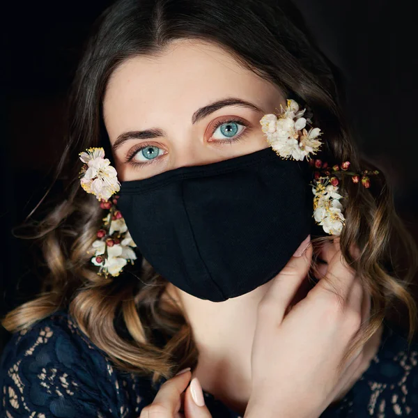 Bahar çiçekleriyle süslenmiş siyah koruyucu maskeli genç ve güzel bir kadın kendini karantinaya almak için evde yalnız kalsın. Ev karantinası, COVID-19 önleme, Coronavirüs salgını durumu