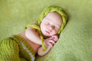 Açık yeşil şapkalı şirin bir bebek ve yeşil arka planda uyuyan bir pantolon.