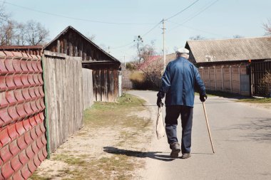 Koruyucu maskeli yaşlı bir adam, eldivenler, eski giysiler içinde, eko çanta ve değnekle köye giden yol boyunca yürüyor. Eski karantina insanları, COVID-19 önleme, Coronavirüs salgını durumu
