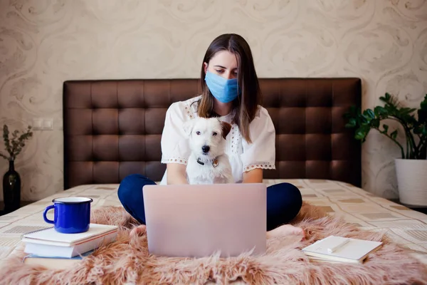 Young woman in protective mask with pet works, studies at laptop at home. Working from home. Concept home quarantine, prevention COVID-19, Coronavirus outbreak situation