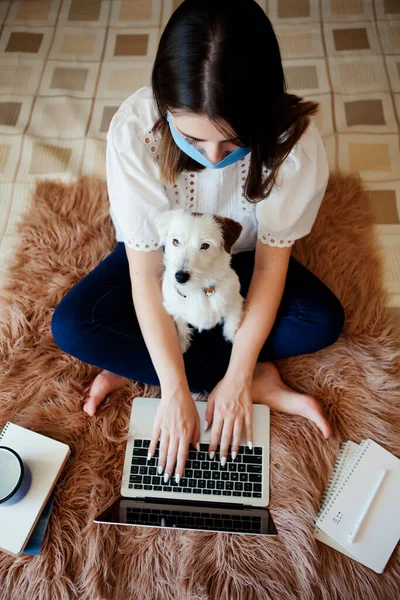 Young woman in protective mask with pet works, studies at laptop at home. Working from home. Concept home quarantine, prevention COVID-19, Coronavirus outbreak situation