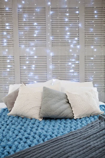 Pillows, knitted cozy blankets on bed on wall with light bulbs garlands background. Trendy room design. Copy space