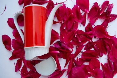 Kırmızı ve beyaz porselen bir kahve demliği, kıçı parçalanmış kırmızı şakayıkların üzerinde duruyor. Yapraklar beyaz bir arka planda yatıyor..