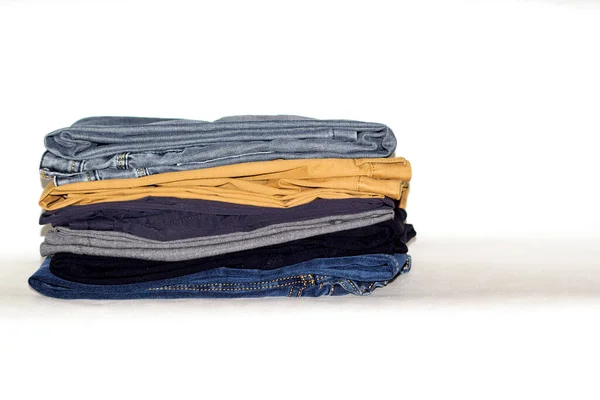 Jeansy o różnych kolorach leżą w stosie na białej powierzchni. Są czarne, brązowe, niebieskie i żółte. — Zdjęcie stockowe