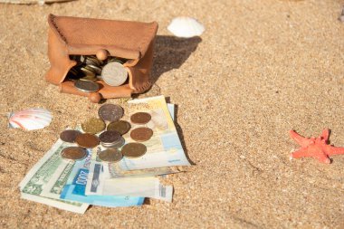 Kağıt banknotlar farklı ülkelerin paralarının altındaki deniz kumu üzerinde yatar. Yakınlarda içi bozuk para dolu çamurdan bir çanta var. Tatil için para biriktirme kavramı, denizde dinlenme..