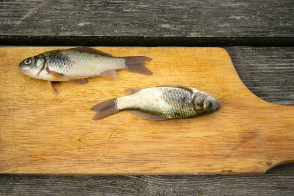 Dois pequenos peixes do rio estão deitados em uma velha tábua de corte quebrada. A placa está em uma mesa de madeira — Fotografia de Stock