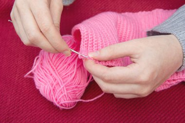 Kadınların elleri, çocuk şapkası için pembe iplik kancası örer. Altında kırmızı bir zemin var. Evde el sanatları.