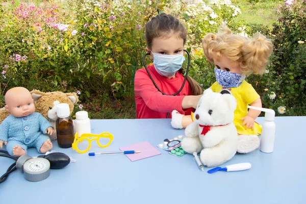 A five-year-old girl in a mask plays a doctor on the street. She listens to the doll.