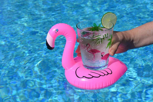 Elinde pembe flamingolarla süslenmiş bir bardak tutan bir kadın, bir dilim limonla köpüren bir su, pembe şişirilebilir flamingo içecek tutacağı içinde yüzme havuzunda.