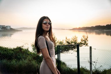 Güneş gözlüklü ve çantalı güzel bir kız. Arka planda güzel bir günbatımı ile nehrin önünde poz veriyor.