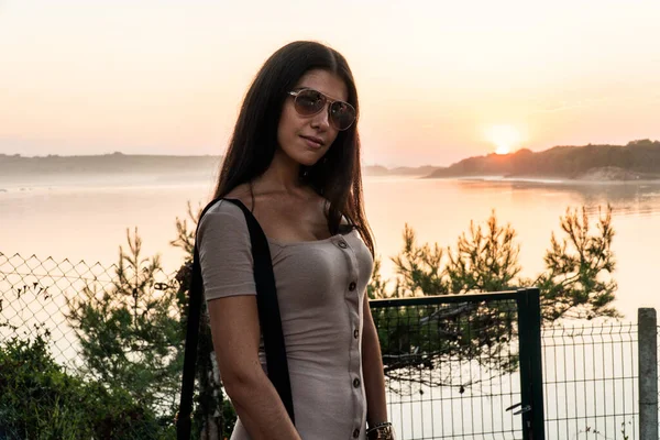 Güzel bir kız, arkasında güzel bir günbatımı olan nehrin önünde poz veriyor.