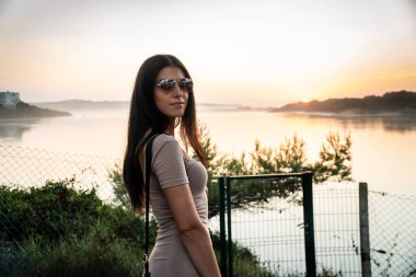 Güzel bir kız, arkasında güzel bir günbatımı olan nehrin önünde poz veriyor.