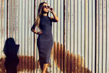 Endüstriyel alanda uzun elbiseli, arka planda paslı çitlerle poz veren genç bir kadın model.