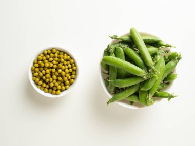 Beyaz bir kasede konserve yeşil bezelyeler ve yanında beyaz bir masada yeşil bezelye kabukları.