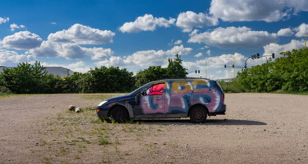 Devastation - a car standing in a closed parking lot, painted in graffiti. in the background blue sky and green trees. Wild parking lot.