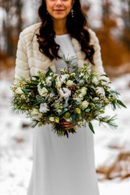 Gelin, kışın, ormanın ortasında, taze karla kaplı bir ormanın ortasında beyaz bir düğün buketi tutuyor.