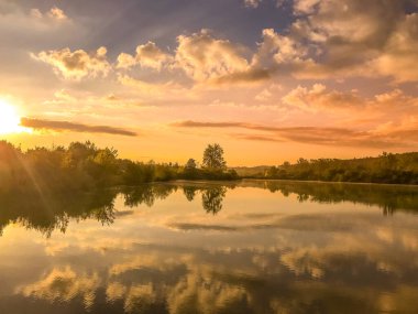 Gün batımında gölde güneş doğarken su kenarında uzanan ağaçların üzerinde ve su yüzeyine yansıyan bulutların üzerinde..