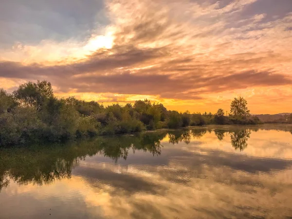 Sunset capture on the pond at the morning sunrise over trees lying near water and moving clouds over water with reflection on water surface.