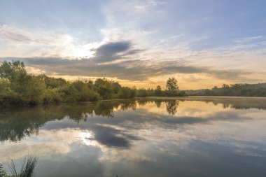 Güneşin doğuşuyla birlikte, suların yakınındaki ağaçların üzerinde uzanan ve su yüzeyine yansıyan bulutların üzerinde hareket eden bir gölet..