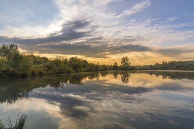 Güneşin doğuşuyla birlikte, suların yakınındaki ağaçların üzerinde uzanan ve su yüzeyine yansıyan bulutların üzerinde hareket eden bir gölet..