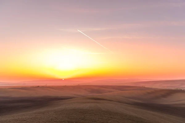 The sun caught during the last moment before setting beyond the horizon with view of a field covered with fog and the farms and fields undulating in the region of South Moravia is also called Tuscany