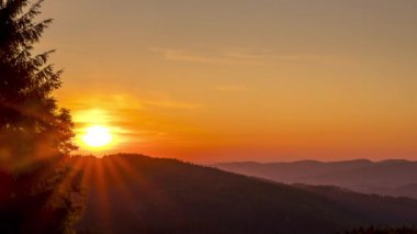 Güneşin zamanı, solunda kozalaklı bir ağaç olan turuncu tepeler üzerinde batarken Beskydy kırsalında arka planda hareket eden bulutlar..