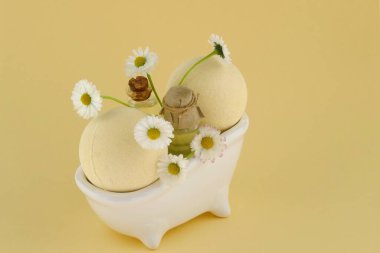 Papatya özlü banyo bombaları. Organik doğal vücut kozmetikleri. Açık sarı arka planda dekoratif beyaz bir banyoda iki banyo bombası topu ve papatya çiçekleri. Banyo için kozmetik..