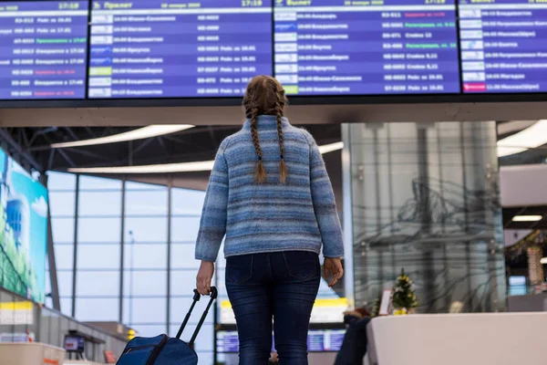 A beautiful young girl with a suitcase in her hand at the airport looks at the flight information Board. The girl is waiting for the departure of the plane.