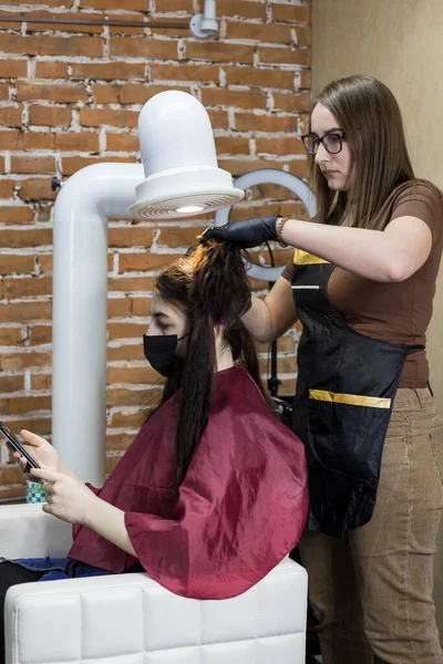 The client-girl with a phone in her hands, sitting in a protective mask and under a hood from the fumes of formaldehyde, while the hairdresser makes her keratin hair strengthening