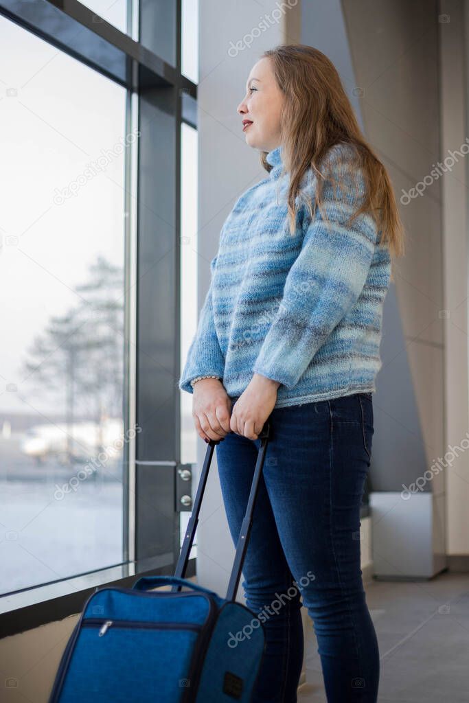 A beautiful tourist with a blue suitcase looks out the window and smiles waiting for the announcement of boarding a plane to fly on vacation. Concept of tourism and travel.
