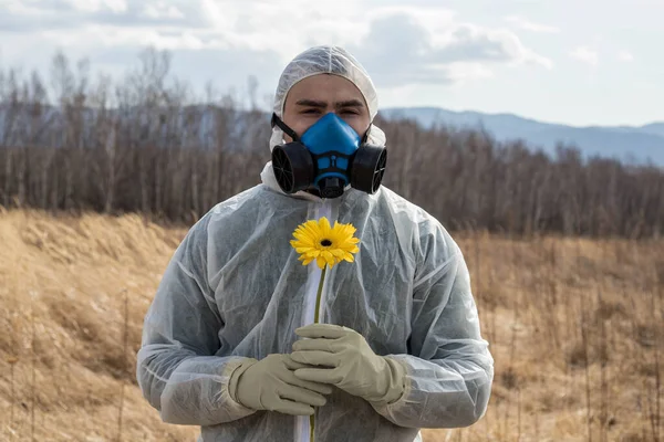 A man in a respirator and chemical protective suit with a yellow flower in his hands. Environmental pollution. Environmental disaster. Allergy to pollen. Highly allergic individual.