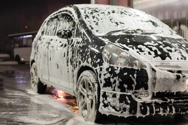 Black dirty car in white soap foam at car wash service station