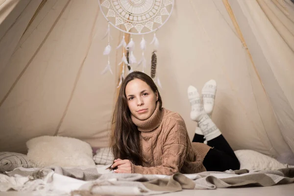 A beautiful young brunette woman in a warm brown sweater is lying in a wigwam. Cozy wigwam with a dream catcher.