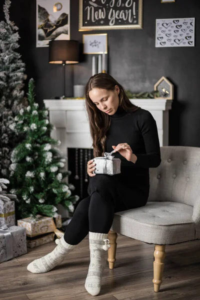 A young woman with a gift is sitting by the fireplace, next to a Christmas tree decorated with toys and garlands. a warm and cozy evening in Christmas interior design. holiday living room. new year.