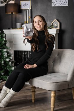 Siyah elbiseli genç bir kadın şöminenin yanında bir Noel ağacının yanında oturuyor ve elinde bir hediye var. Genç bir kadın hediyeyle mutlu olur. Noel zamanı. Şenlikli atmosfer.