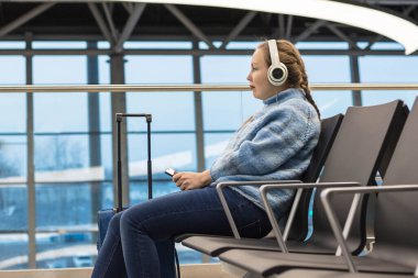 Kulaklık takan bir kadın havaalanı salonunda oturmuş uçuşunu bekliyor..