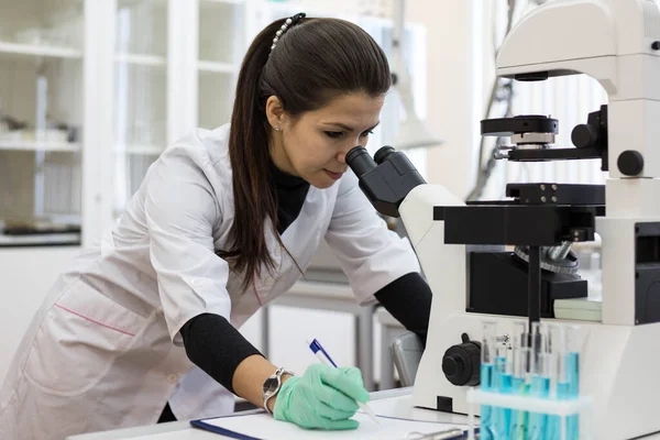 A female chemist looks through a microscope and makes notes in a journal. Scientific research using a microscope in the laboratory. Woman scientist.