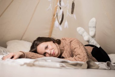 A beautiful young brunette woman in a brown sweater lies smiling in a cozy wigwam clipart