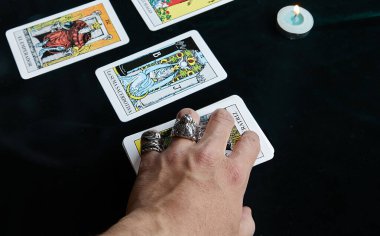 Yüzüklü bir el, tüten bir mumun yanındaki siyah bir kumaşa tarot kartları koyar.
