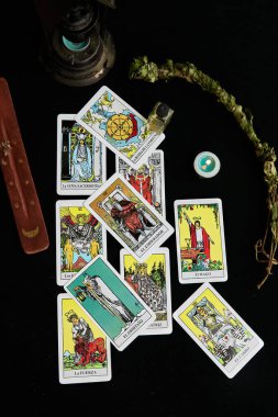 Koyu renk kumaş üzerinde tarot kartları, esans şişesi, adaçayı dalı, mum tütsü kahverengi masa altın ay ve altın yıldızlı.