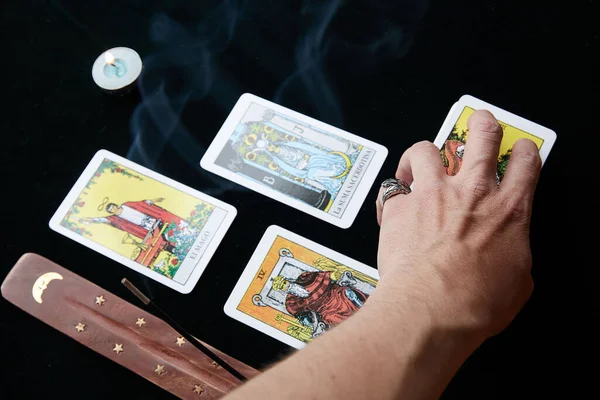 Yüzüklü bir el, tüten bir mumun yanındaki siyah bir kumaşa tarot kartları koyar.