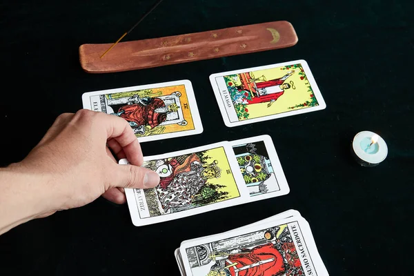 Bir el, kandilin yanındaki koyu bir kumaş üzerinde tarot kartları ve altın ay ve altın yıldızlı kahverengi bir masada tütsü alır.