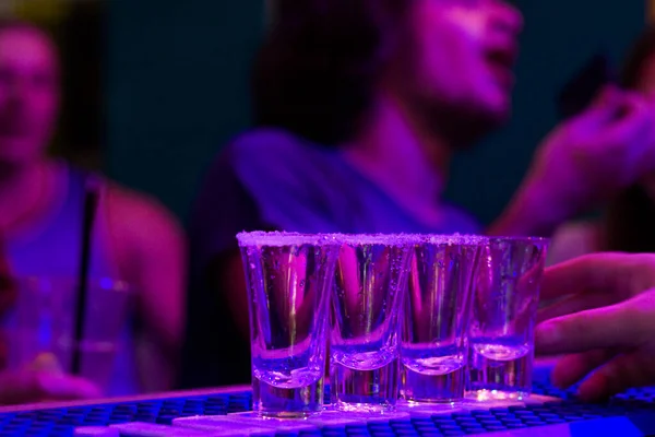 Tequila shots on the bar table at night party, people on the background, alcoholic full shots with blue led light in Tbilisi night club.