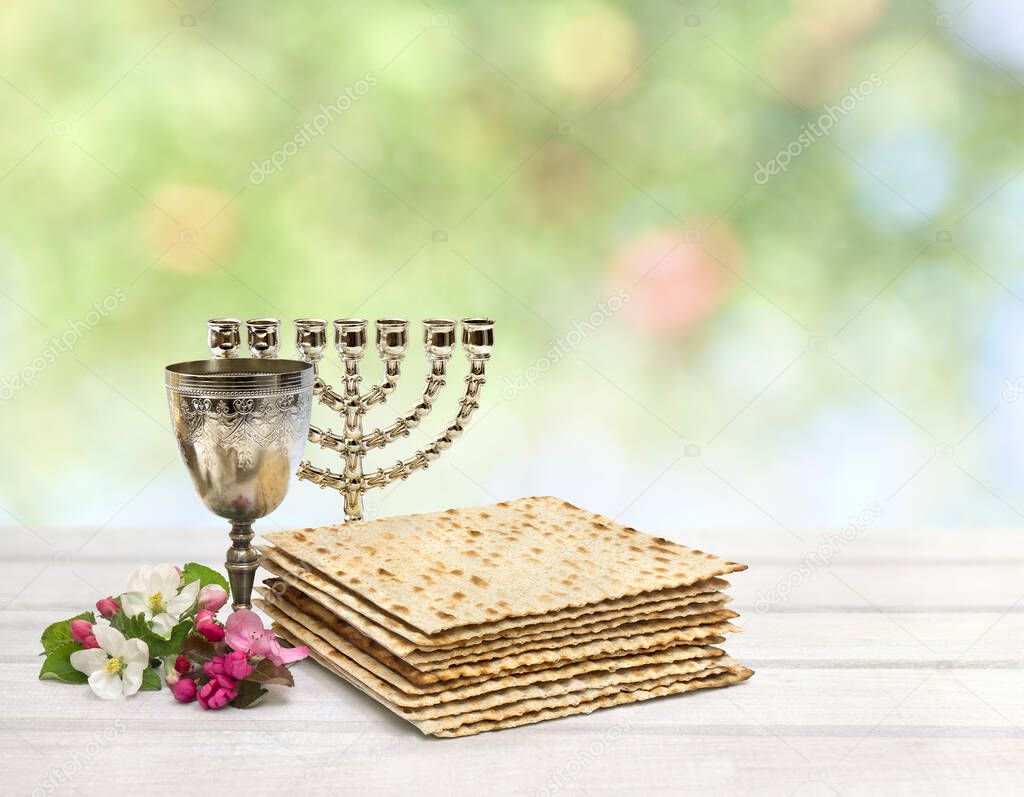 Matzo, wine, menorah and pink flowers apple tree for passover celebration on white plank table on blur nature background with space for text.