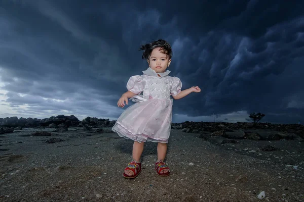 A baby girl are standing by the stormy sea and looks into the camera.